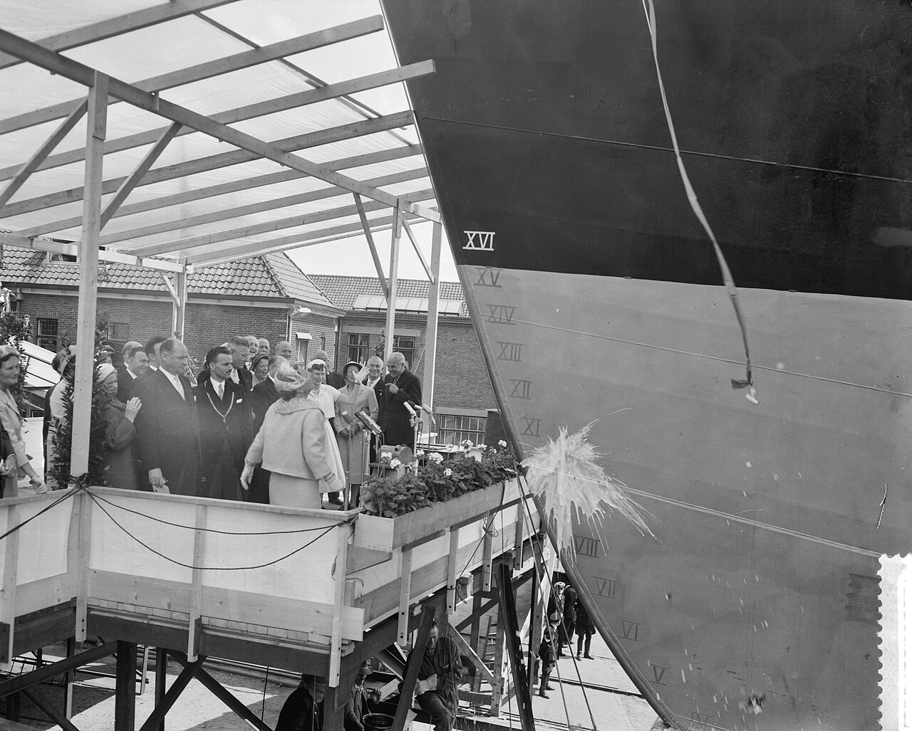 A black-and-white photograph of a woman in a light coat breaking a bottle against the bow of a large ship on a shipyard slipway, watched by a group of dignitaries on a viewing platform.